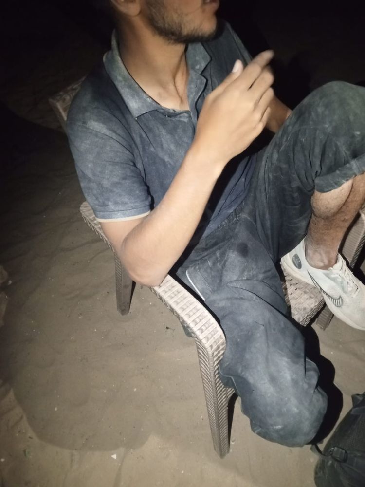 Abdullah after returning home from the GHF aid site in Rafah. He sits on a plastic chair with one knee up. His clothes are covered in dust.