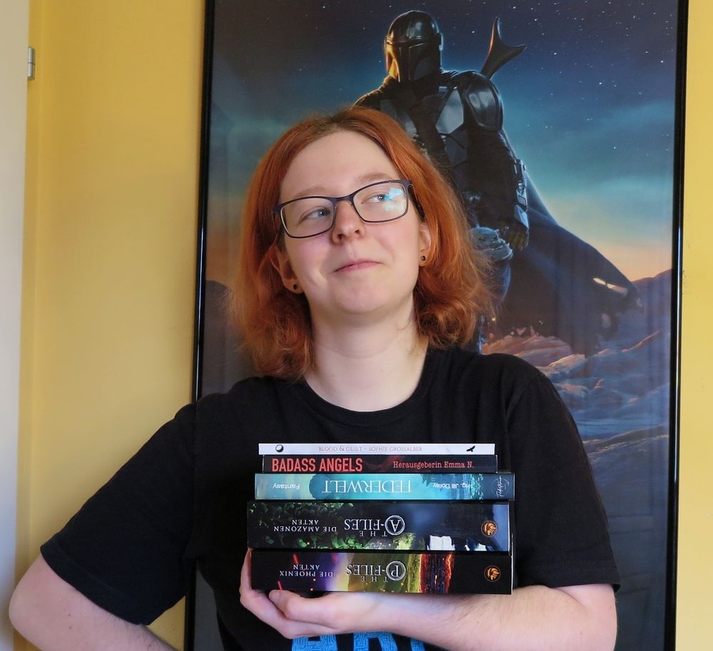 Picture of afab person with glasses and orange, shoulder-length hair holding a stack of books.