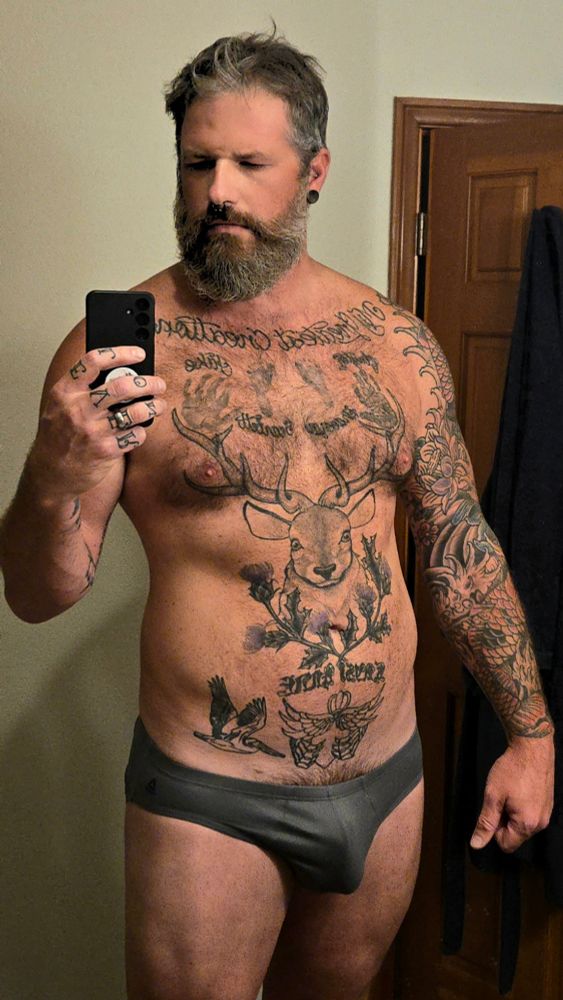 A muscular, bearded man with short, slightly messy salt-and-pepper hair is taking a mirror selfie in a bathroom. He is standing shirtless, wearing only gray briefs. His expression is neutral, with his eyes slightly squinted as he looks at the phone in his hand. He has a septum piercing, stretched earlobes with black plugs, and several rings on his fingers.

His chest, stomach, and both arms are heavily tattooed. A large deer head with antlers covers most of his chest and upper abdomen, surrounded by additional script and decorative designs. His right arm features a full sleeve tattoo, while his left arm has multiple smaller tattoos. The background shows a beige wall, a wooden door, and a dark towel hanging on a hook. The lighting is warm and soft, typical of indoor bathroom lighting.
