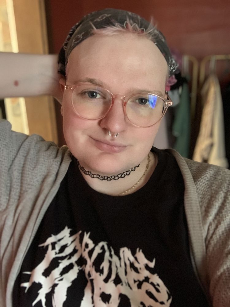 close up selfie of kaycie, a white trans woman. im wearing a black bandana with tufts of pink hair poking out, pink clear round glasses, a black shirt with metal band text that reads "my bones hurt" and a gray sweater. I'm smiling mischievously and have a hand behind my head. my silvery blue eyes look really beautiful catching the light.