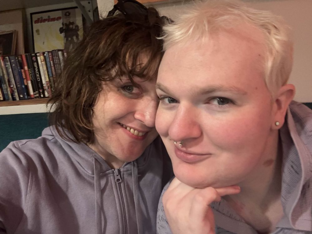 a closeup of two beautiful white trans women wearing purple sweatshirts. on the left is sky, with shoulder length slightly curly mousy brown hair. she leans into and slightly behind kaycie, who has short bleached hair. they are smiling so genuinely and deeply 