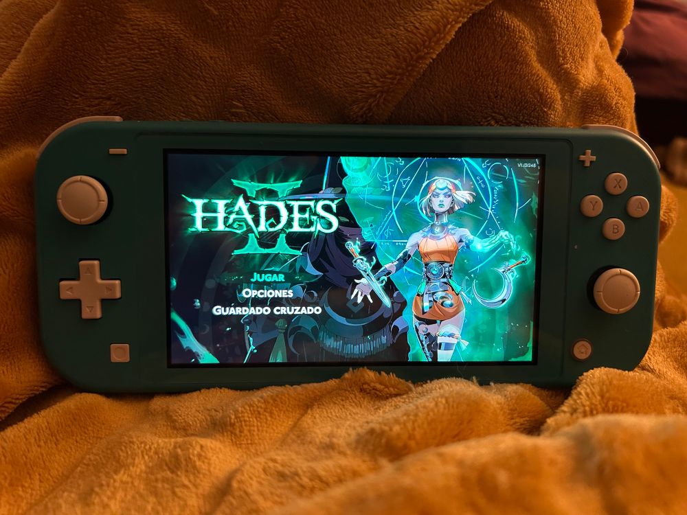 Switch Lite turquesa sobre una manta amarilla y con el menú principal de Hades 2 en pantalla.