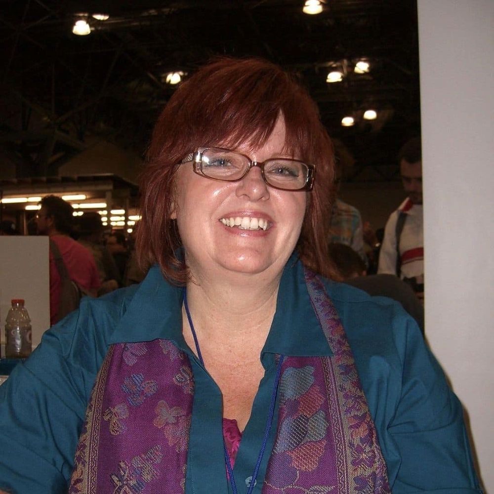 Gail Simone, a white woman with shoulder length straight red hair, wearing rectangular glasses,  a teal shirt and a purple scarf.  Definitely not a bear (this is a joke among Gail's fans)