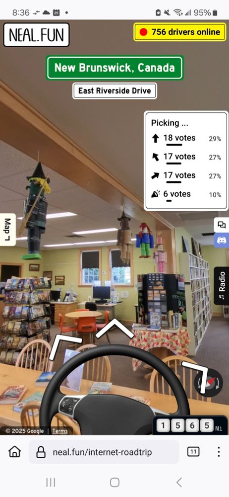 Interior of a library with yellow walls, shelves of books, tables & chairs, & various doll figures hanging from the ceiling beams. A car steering wheel is superimposed over top. 