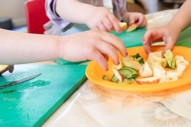 Children’s hands reaching for pieces of sliced fruit and cucumber on a yellow plate, with a green cutting board and a knife visible on the table.
Photo credit: Dean Leivers