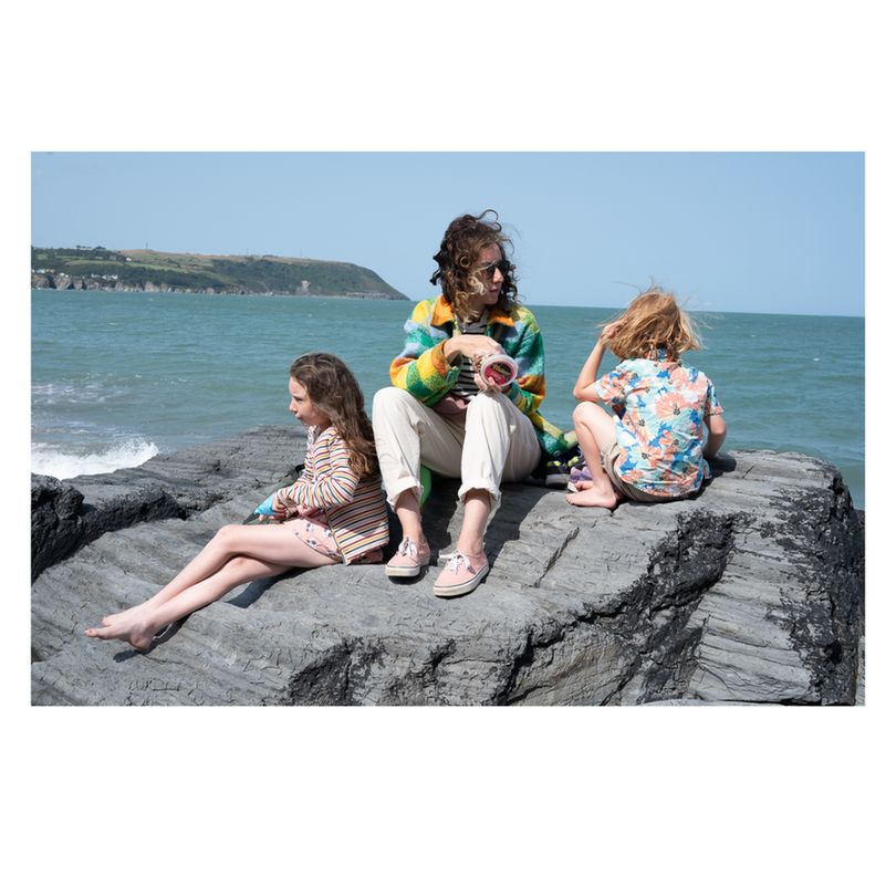 Three people sit together on a large rock by the seaside, facing the ocean. The person in the center wears a colorful striped sweater and light trousers, holding something in their hands. The person on the left wears a striped long-sleeve shirt and shorts, while the one on the right wears a floral shirt and shorts. Behind them, waves roll in and a distant coastline is visible under a clear sky.