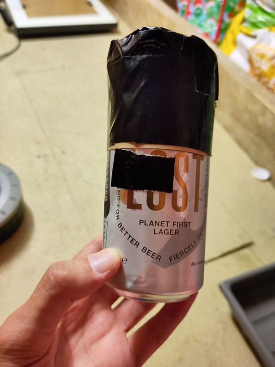 A hand holds a homemade pinhole camera made from a recycled beer can labeled "Lost Planet First Lager." The top of the can is covered with black tape, and a rectangular piece of tape likely conceals the pinhole opening used for capturing the image.