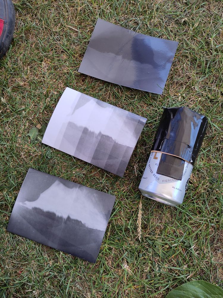 A scene showing three black-and-white photographic prints laid out on grass alongside a makeshift pinhole camera made from a silver beer can covered in black tape. The prints depict landscape scenes with varying contrasts, likely results from a darkroom photography workshop. A person's shoe is partially visible in the top left corner.