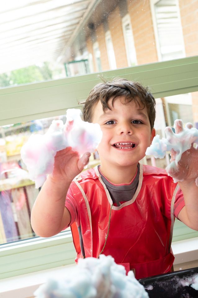 A young child wearing a red apron is engaged in a sensory play activity, holding up hands covered in colorful foam, standing indoors by a window. Photo credit: Dean Leivers