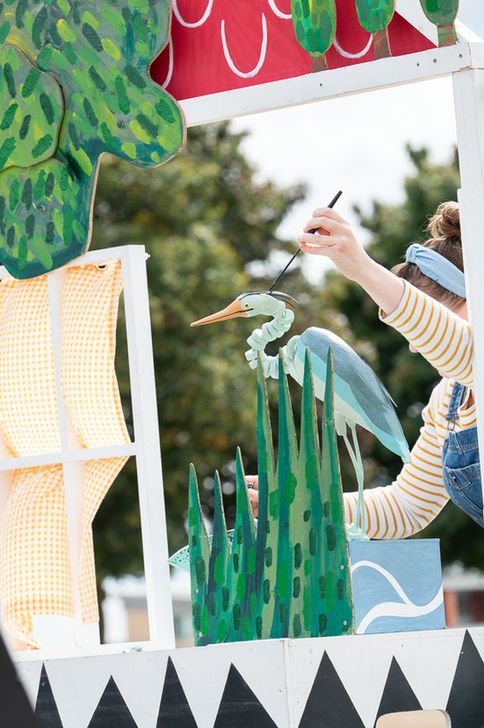 A person in striped sleeves and denim overalls operates a bird puppet resembling a heron or egret using a black rod. The puppet is set among painted green reeds and stylized water waves, evoking a wetland scene. Surrounding props include a red and green watermelon-like structure, yellow checkered curtains, and lush green foliage. The backdrop features trees and a cloudy sky, indicating an outdoor setting. The scene is colorful and imaginative, showcasing detailed craftsmanship in both puppet and set design.