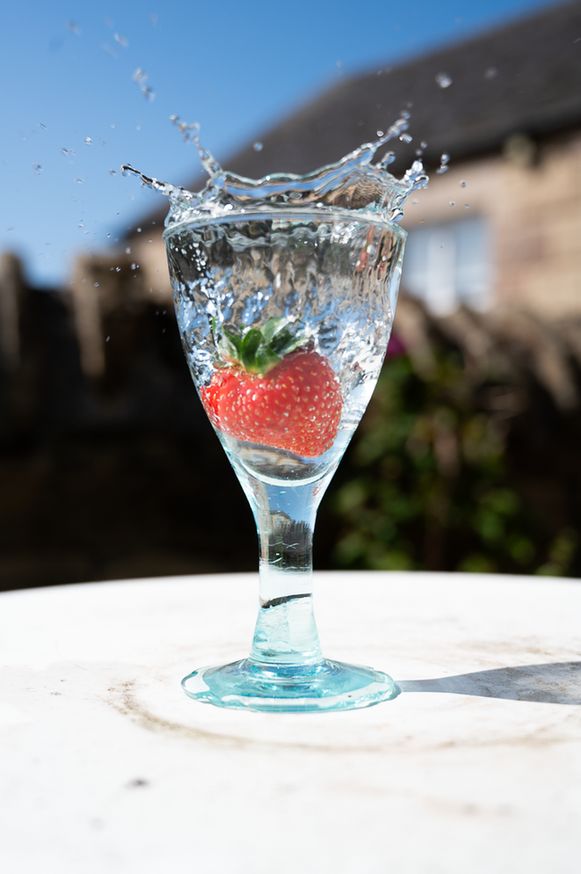 A fresh strawberry splashes into a clear glass of water, captured mid-motion with droplets flying outward. The glass sits on a white surface outdoors, with a blurred background of buildings and blue sky.

