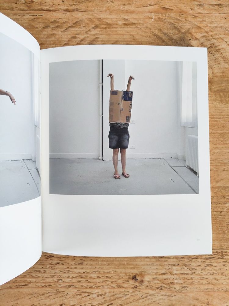 A person stands in a sparse white room with a cardboard box covering their head and upper body. Their arms are raised above the box, and they are wearing shorts and sandals. The scene is playful and absurd.
Photo Credit: Sian Bonnell