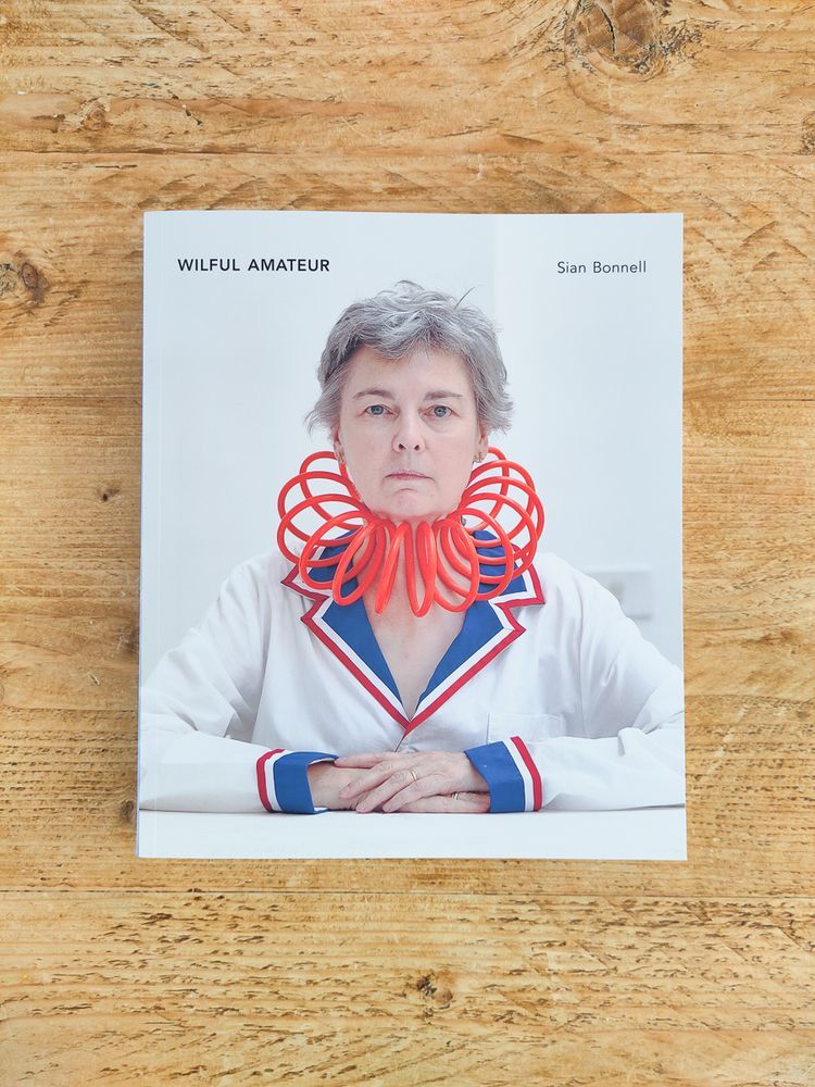 A book cover titled "Wilful Amateur" by Sian Bonnell. The cover image features a serious-looking person in a white outfit with red and blue trim, wearing a large, whimsical red plastic spiral collar around their neck. The background is a minimalist white space.
Photo Credit: Sian Bonnell