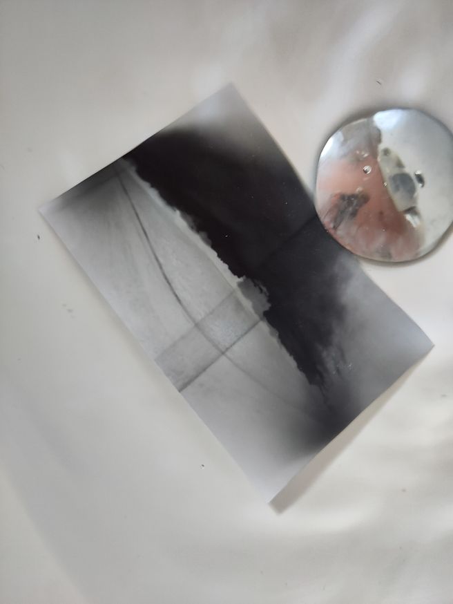 A black and white photographic print soaking in a white tray filled with liquid. The image appears to show a landscape or road scene, partially developed, with a round, shiny metal object (possibly a weight or tool) resting beside it.