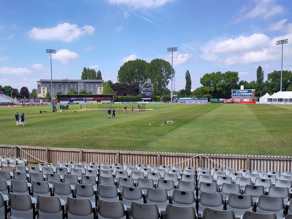 A view of a cricket ground from the stands, showing empty grey seats in the foreground, a green pitch with a few people scattered across it, and buildings, trees, and floodlights in the background under a partly cloudy blue sky.