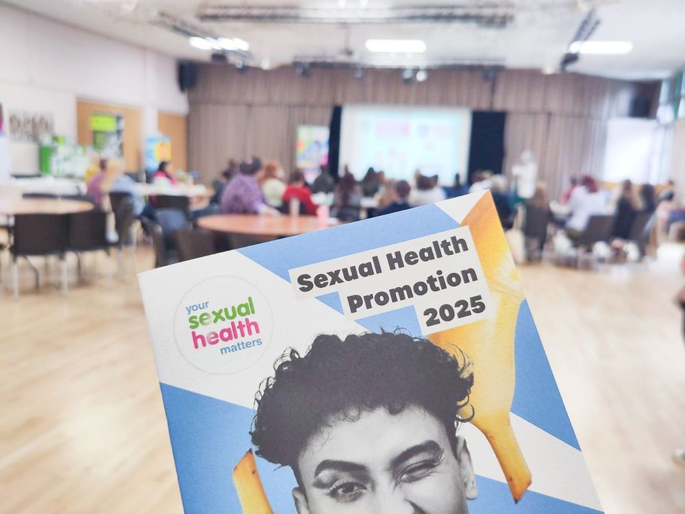 A hand holding a leaflet titled "Sexual Health Promotion 2025" with a colourful design, in a large room where people are sitting at round tables facing a presentation screen and speaker at the front.
