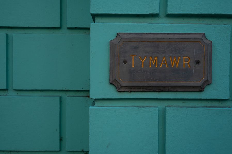 A close-up of a dark plaque with gold lettering that reads "TYMAWR," mounted on a teal-painted brick wall. The sign is slightly weathered, and the wall has a textured, geometric pattern.