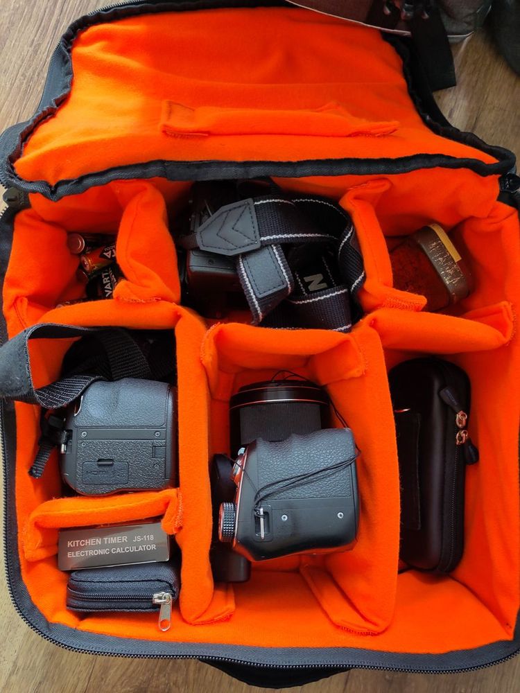 Open camera bag with bright orange padded compartments containing multiple DSLR cameras, camera accessories, a lens, batteries, and a small electronic timer.