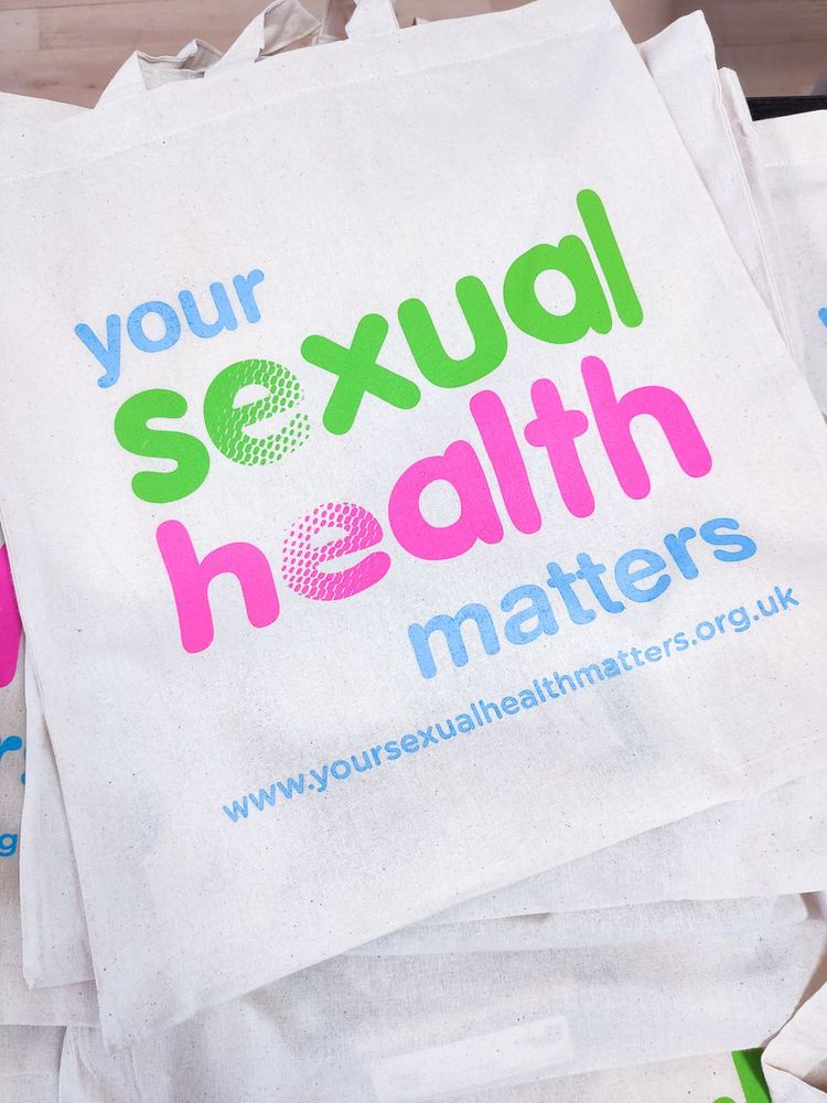 A close-up of a white tote bag with colourful text that reads "your sexual health matters" and a website address "www.yoursexualhealthmatters.org.uk" printed in blue at the bottom.