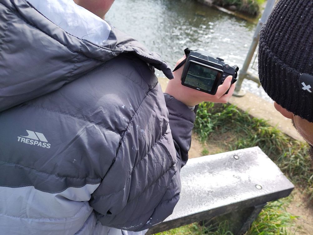 Two people standing by a riverside path; one is holding a digital camera and reviewing a photo on its screen, wearing a grey Trespass puffer jacket.