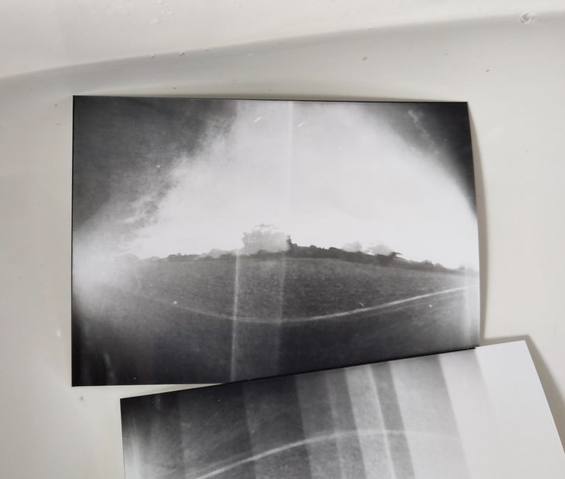 A black and white photograph, possibly a pinhole camera image, shows a landscape with a horizon line and some trees or distant structures. The image features bright and dark areas with visible light leaks or exposure bands, giving it a hazy, experimental look. Part of another similarly styled photo with vertical light streaks and exposure marks is partially visible underneath. Both photos rest on a white surface, suggesting a darkroom or photographic workspace.