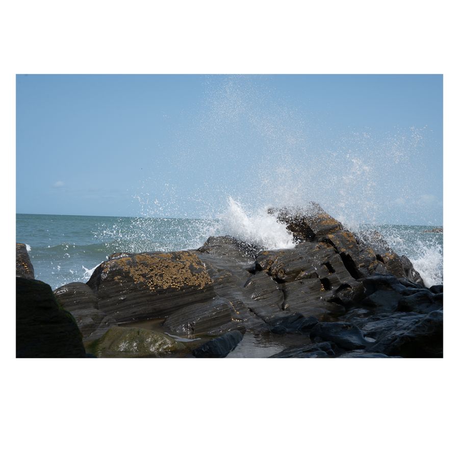 Waves crash energetically against large, dark rocks along a rugged shoreline. Some rocks are speckled with barnacles or marine growth, adding texture. The ocean behind is calm under a clear blue sky, while the splashing water in the foreground captures the dynamic force of the sea.