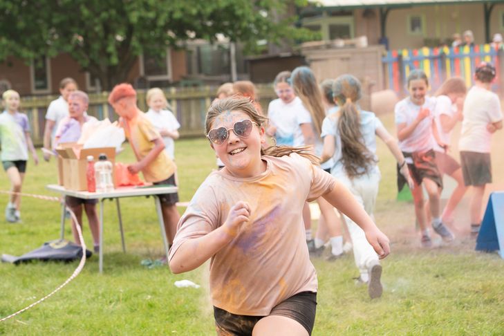 A girl wearing heart-shaped sunglasses runs toward the camera, smiling, during a vibrant outdoor color run surrounded by other children covered in colorful powder.