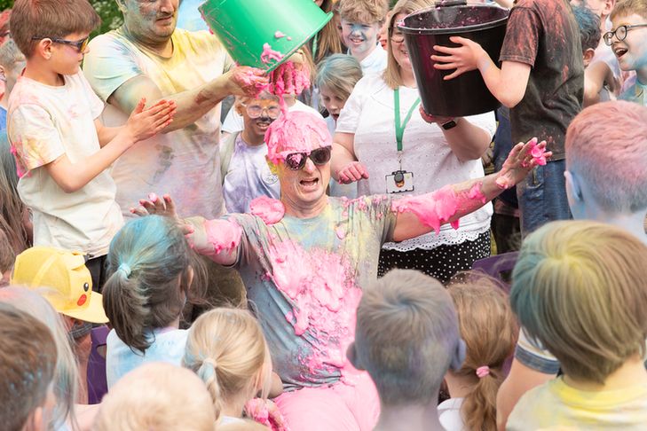 A group of children and adults cheer as a man sitting on the ground is doused in bright pink foam, laughing and covered in colorful powder during a festive school color run event.