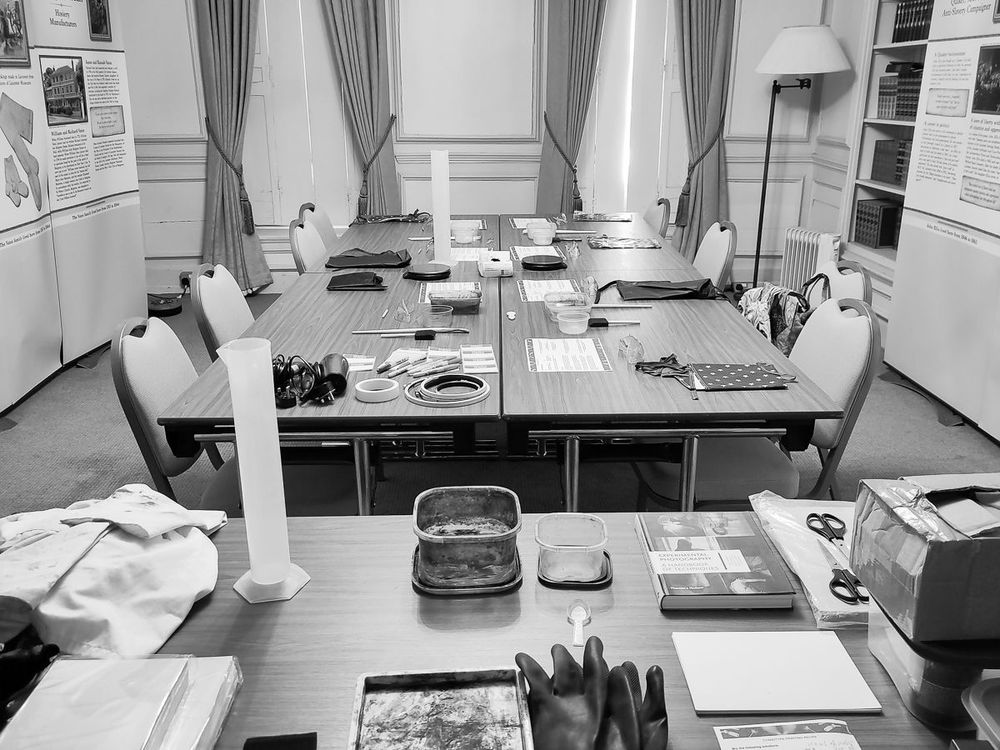 A black and white photo of a classroom or workshop setup, with several tables arranged in a U-shape, surrounded by chairs. The tables are covered with various materials and equipment, including gloves, measuring cylinders, trays, scissors, books, and boxes. Informational display boards and curtains are visible in the background, suggesting a historical or educational setting.