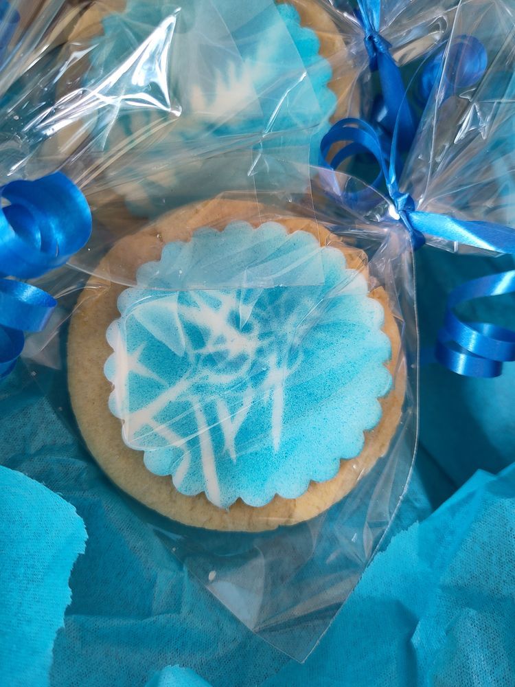 A close-up of round sugar cookies wrapped in clear plastic with blue ribbons. The cookies have a scalloped edge white icing base with blue airbrushed patterns resembling snowflakes or abstract designs. They are placed on a blue fabric or tissue background.