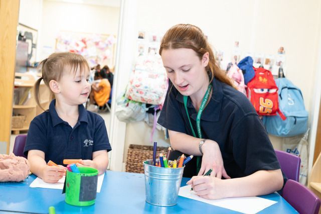 A young girl sits at a table holding markers while a teacher with a Greenfield Primary School lanyard helps her with a drawing activity.
Photo credit: Dean Leivers