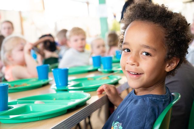 A smiling young boy sits at a lunch table with green trays and blue cups, surrounded by other children in a school cafeteria.
Photo credit: Dean Leivers