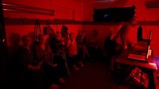 A group of people are sitting along a wall in a dark room illuminated by red light, commonly used in photographic darkrooms. One person is standing at a table with trays and photographic equipment, suggesting a workshop setting.