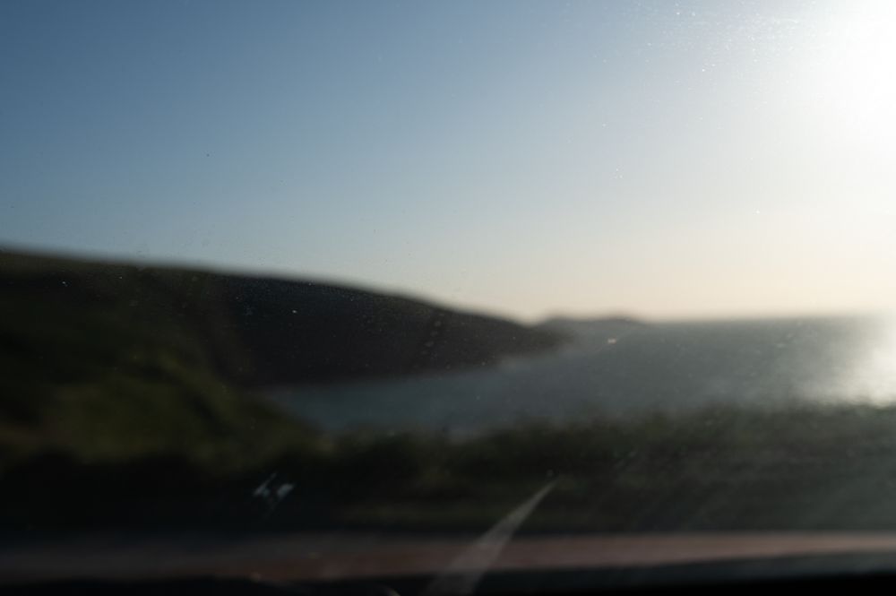 A blurry view of a coastal landscape taken through a dirty car windshield. The photo shows a bright sky and sunlight reflecting off the ocean, with grassy cliffs in the distance. The windshield has visible smudges and dust particles, partially obscuring the view.