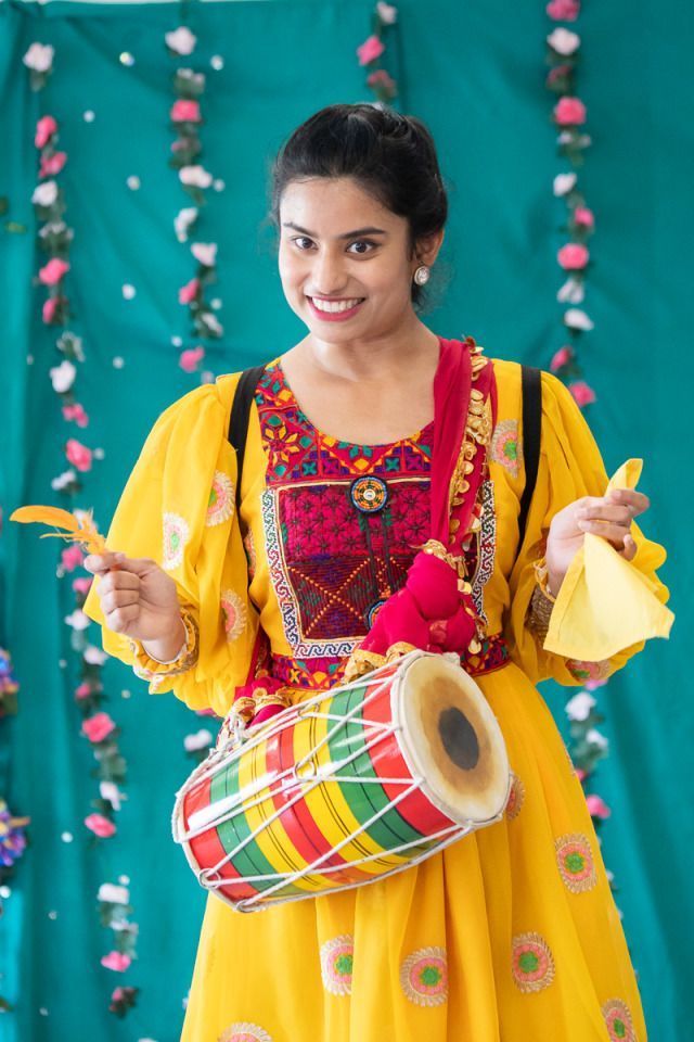 A person wearing a bright yellow traditional dress with colorful embroidery and floral patterns stands in front of a teal backdrop decorated with hanging flowers. They hold a small decorated drum (dhol) strapped across their body and yellow cloth in one hand, with a feather in the other, suggesting participation in a cultural or folk performance.