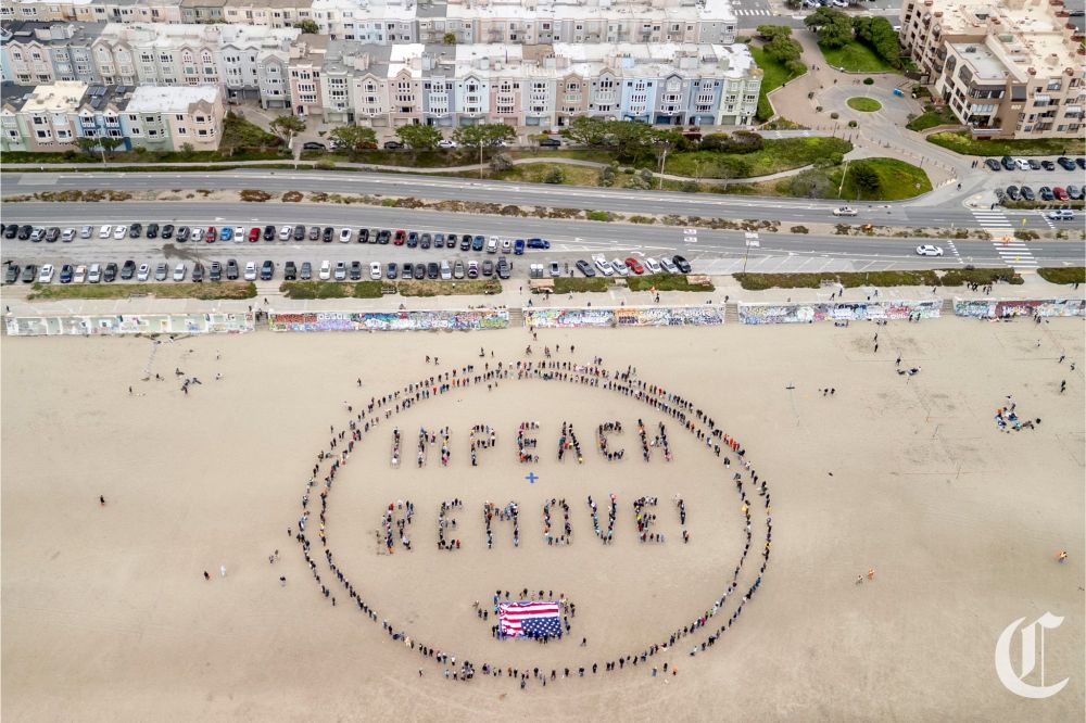 Drone photo of Ocean Beach in San Francisco with a group of people gathered in a circle, forming the words "impeach + remove" inside. An upside-down American flag is inside the circle as well. 