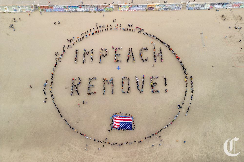 Drone photo of Ocean Beach in San Francisco with a group of people gathered in a circle, forming the words "impeach + remove" inside. An upside-down American flag is inside the circle as well. 