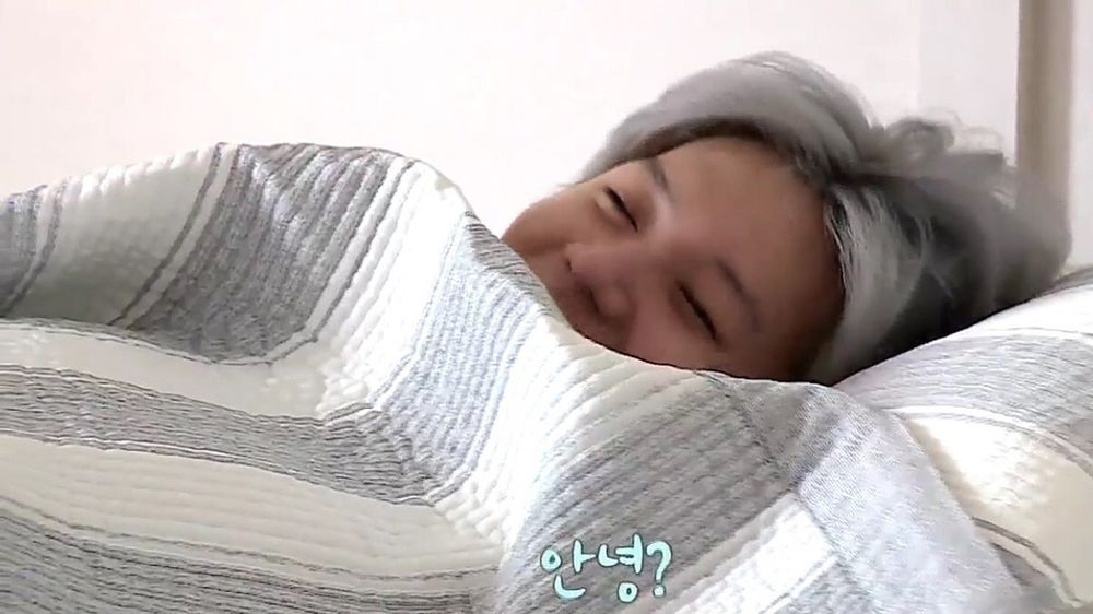 Sleepy, smiley, Taemin under the covers, silver hair poofed over his pillow, eyes mostly closed but clearly smiling. Caption says “안녕?“ Indeed.