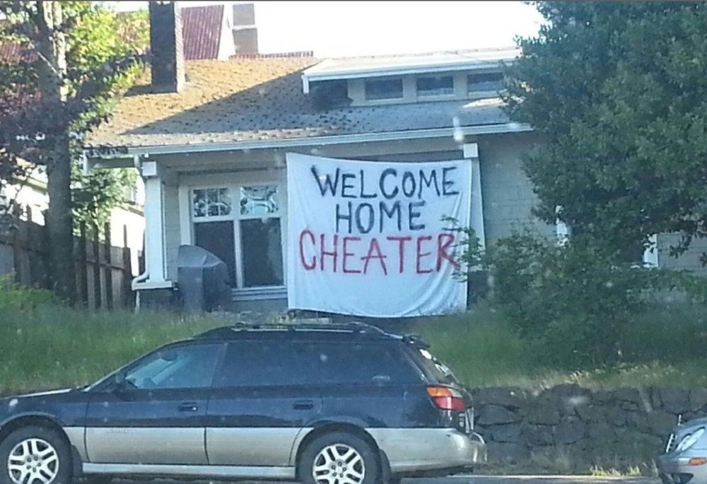 A house with a large cloth hung in front of a house, messily spray-painted with the words "WELCOME HOME CHEATER."