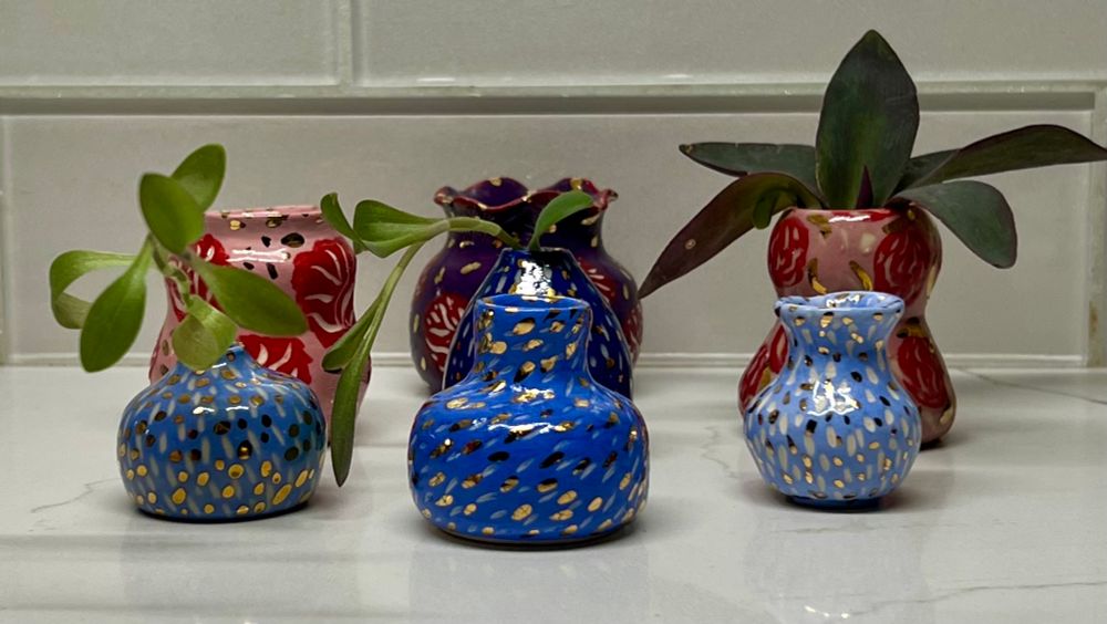 Set of seven miniature vases a various shapes and heights, featuring sgraffito abstract design, in shades of blues, pinks, red and purple gloss glazes, all finished with 22 karat gold metallic glaze accents. 