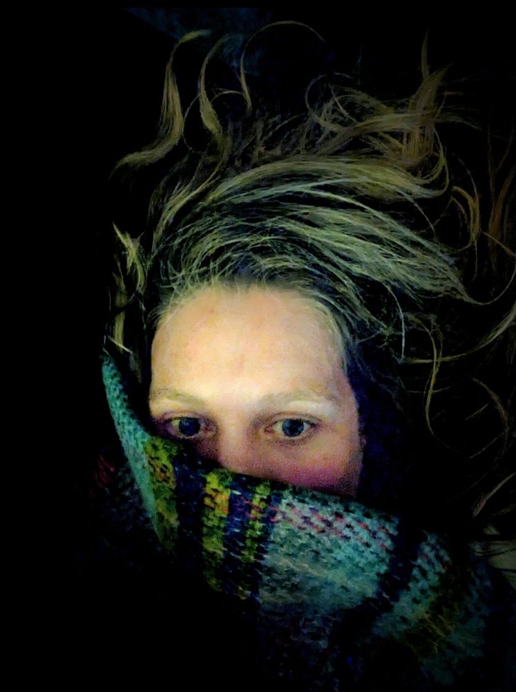 A colour picture of a partially covered face in a blue wooden blanket there are lines of green and dark blue too. The only areas of the face showing are the eyes and forehead. The mouth and nose are covered. The hair is splashed across the dark background. 