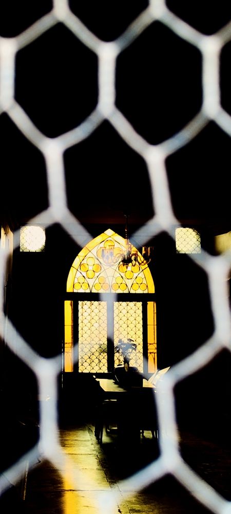 A colour photograph of an arched window which is glowing a yellow colour. There is a dark cross in front and a table and chairs  the surround of the window is dark apart from a light glow on the table.
There is wire mesh covering the picture which is present but not obscuring. 