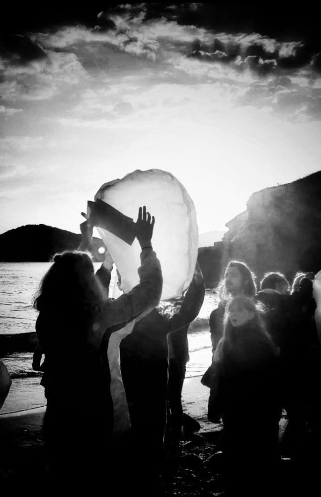 A b&w photograph of a group of people on a beach with the main person having their back to me holding up a lantern ready to go up in the air. There are other people facing me but they're focused on the lantern. In the background is the sea and some cliffs. There is a sad story to this. But it's not mine to tell. 