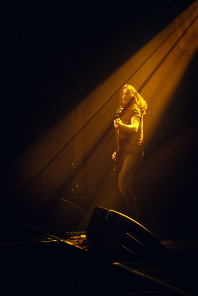 A guitarist stands on stage in a dramatic beam of golden light, playing to the audience during a tribute rock concert.