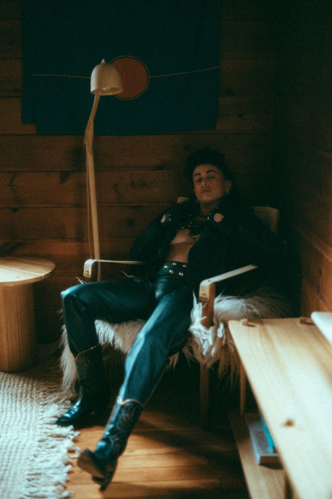 Devorah seductively lounges on a chair in leather boots, chaps,  jacket and harness. He pulls the jacket open.