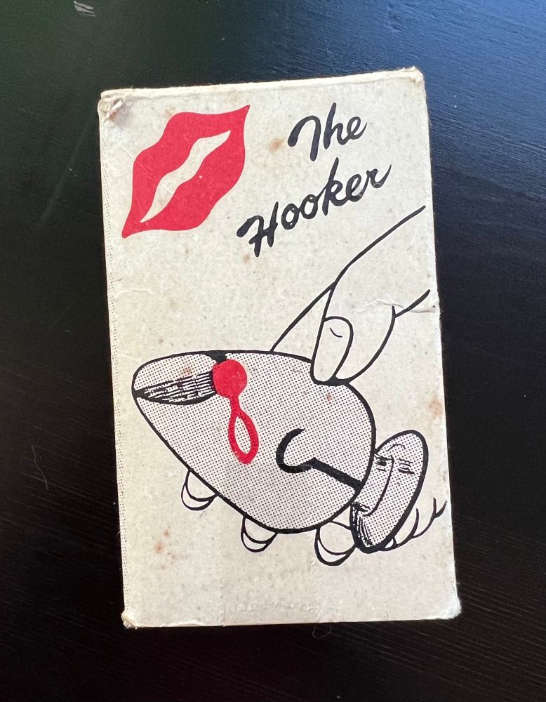 A cardboard box depicts an illustration of hand holding a glass butt plug. It’s a water ring toy with a black fishing hook and round red ring bait inside. It says “The Hooker” and features an image of red lips.