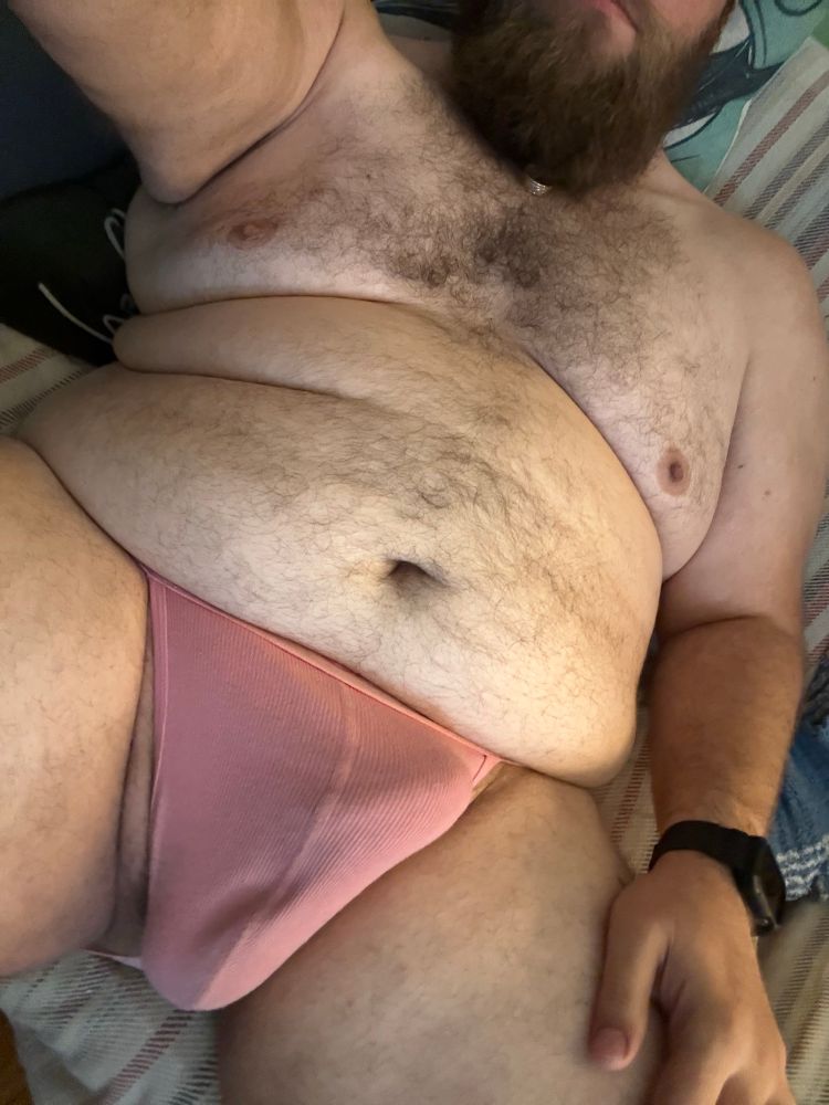 A body focused shot of Husky laying back on the couch in a light pink nasty pig jocks. His hairy belly and chest are prominently on display, and there’s a conspicuous bulge tenting his jock