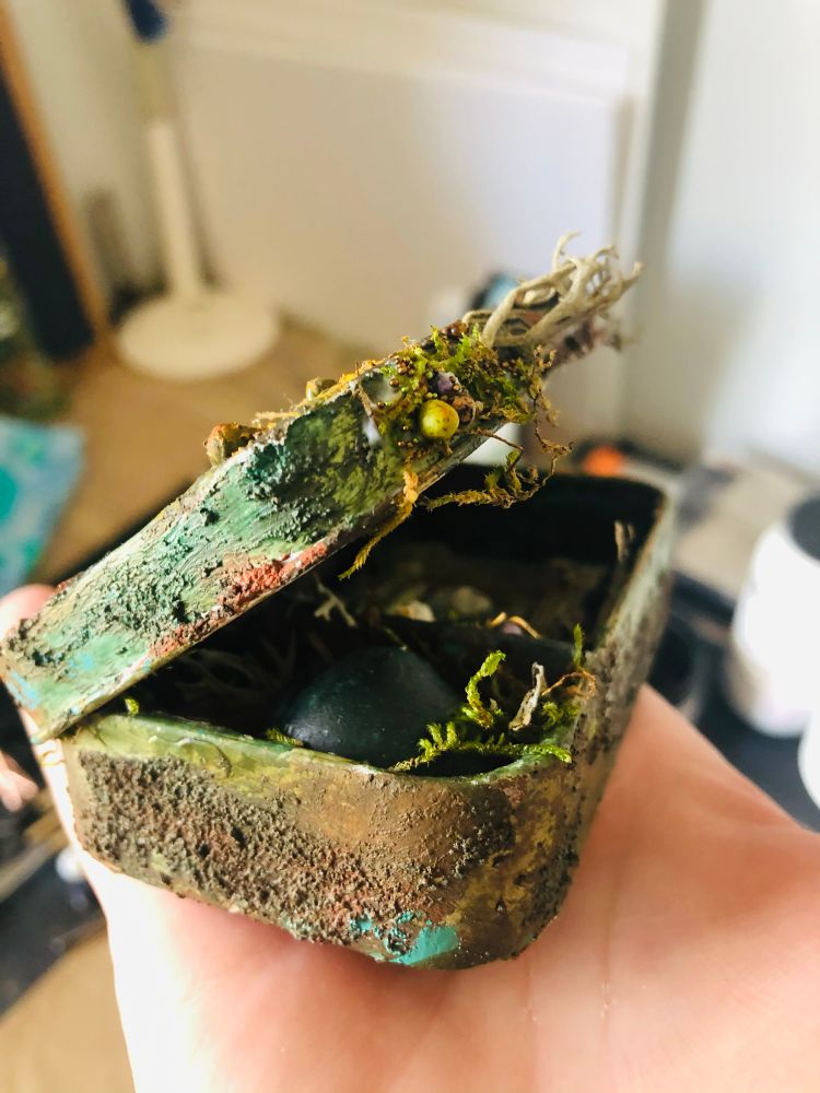 The outside of the altered Altoid tin. It has been painted, textured, and aged to look like it was lying on the forest floor for years. Small Altoid tin resting, half open on my right hand