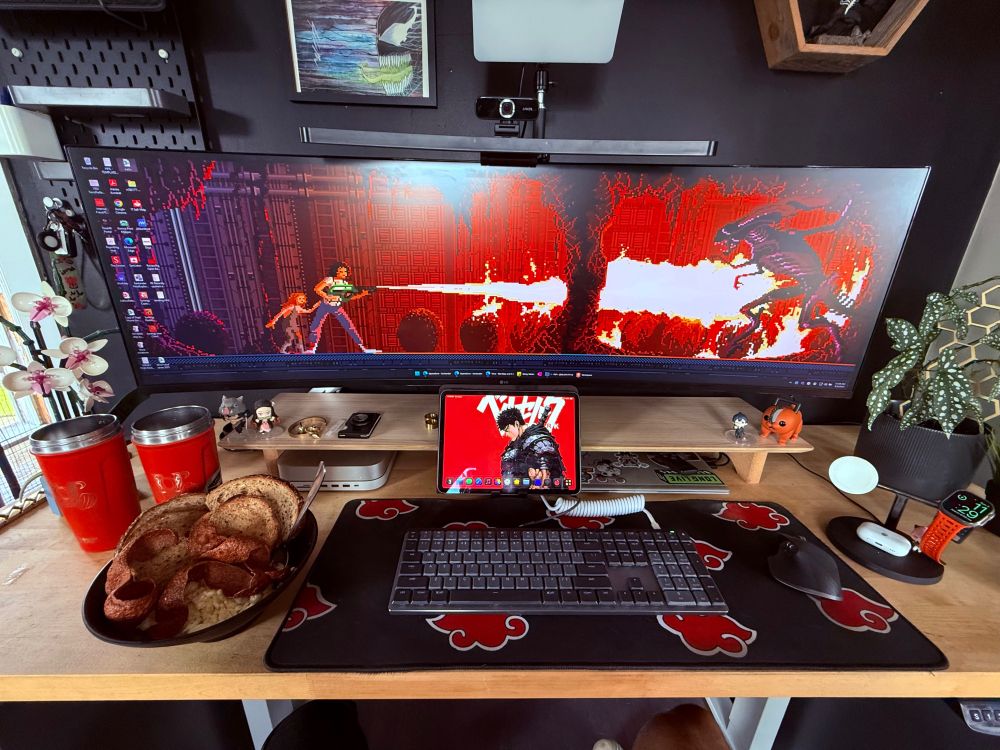 This is a well-organized, anime-themed computer setup with a cozy and personal vibe. Here’s a breakdown of what’s visible in the image:

Desk & Tech:
	•	Monitor: Ultra-wide curved monitor showing a pixel-art action game or animation with a red/fiery color theme.
	•	Tablet: A tablet (likely an iPad) sits below the monitor, docked with a red Berserk wallpaper featuring Guts.
	•	Keyboard & Mouse: Wireless keyboard and ergonomic mouse on a black mousepad with the Akatsuki cloud symbol from Naruto.
	•	Lighting & Webcam: A light bar and webcam are mounted on the monitor for video calls or streaming.
	•	Monitor Riser Shelf: Holds a couple of figures, a charger with Apple Watch and AirPods, and some other accessories.

Personal Touches:
	•	Figures: Anime or gaming-themed mini-figures placed on both sides of the monitor and shelf.
	•	Desk Plant: A polka dot begonia in a black pot on the right.
	•	Wall Decor: Art above the monitor and a wooden hexagonal shelf on the upper right with a small figure inside.
	•	Akatsuki Theme: The mousepad and cups both feature the Akatsuki cloud from Naruto, showing a clear fandom.
	•	Clock & Fitness Band: Digital clock shows 2:57 and a red fitness band (possibly Apple Watch) is charging.

Food:
	•	Meal: A hearty bowl of eggs, turkey bacon and multigrain toast.
	•	Drinkware: Two red tumblers with Gym logo TOP strength project engraved on them.

Extras:
	•	Orchid: Artificial orchid flower for added aesthetics on the left.
	•	Clean Layout: Cable management is tidy, and everything has a place.

This setup combines functionality, anime fandom, and a personalized aesthetic—perfect for productivity, gaming, or content creation.