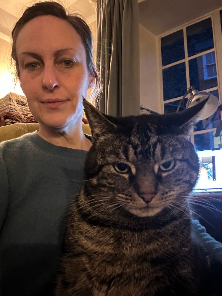 Polly in dim afternoon room with jumper with large cross looking tabby cat siting upright on her knee. Window behind her shows it is rainy and almost dark 